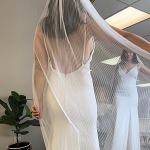 May include: A white wedding veil with a pleated design. The veil is draped over a white wedding dress.