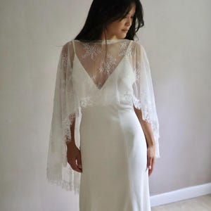 May include: A white floor-length dress with a V-neckline and thin straps, paired with a sheer white lace capelet featuring a delicate floral pattern that drapes over the shoulders and upper chest, creating a layered look with a flowing silhouette.