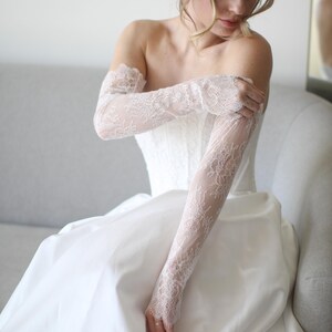 May include: A white wedding dress with a strapless bodice and a flowing skirt. The bride is wearing long, delicate lace gloves. The gloves and dress are a matching white. The bride is sitting on a gray couch.