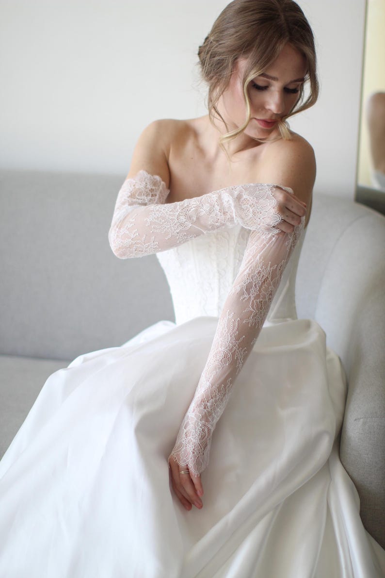 May include: A white wedding dress with a strapless bodice and a flowing skirt. The bride is wearing long, delicate lace gloves. The gloves and dress are a matching white. The bride is sitting on a gray couch.