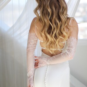 May include: A white bridal gown with a low back and spaghetti straps. The dress features a row of buttons down the back. Delicate, sheer lace sleeves in a light pink hue add a touch of elegance to the ensemble.