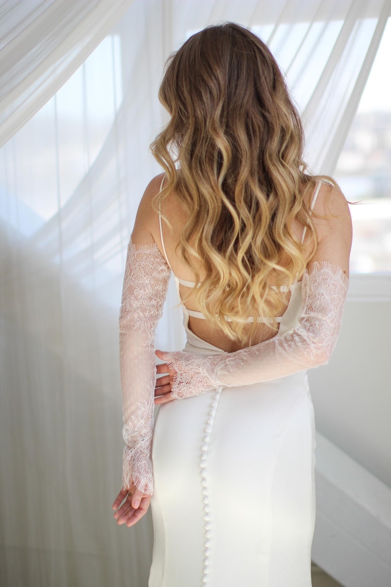 May include: A white bridal gown with a low back and spaghetti straps. The dress features a row of buttons down the back. Delicate, sheer lace sleeves in a light pink hue add a touch of elegance to the ensemble.