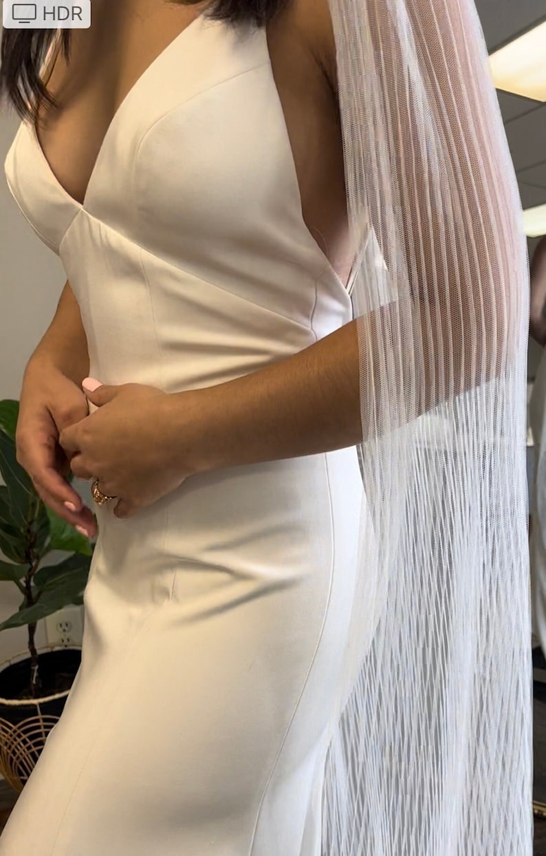 May include: A white sleeveless wedding dress with a plunging neckline and a sheer white cape with a pleated design.
