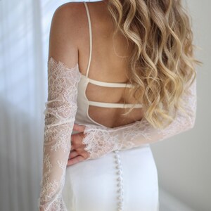 May include: A white wedding dress with a low back and spaghetti straps. The dress features a row of buttons down the back and sheer lace sleeves. The bride's hair is long and wavy.
