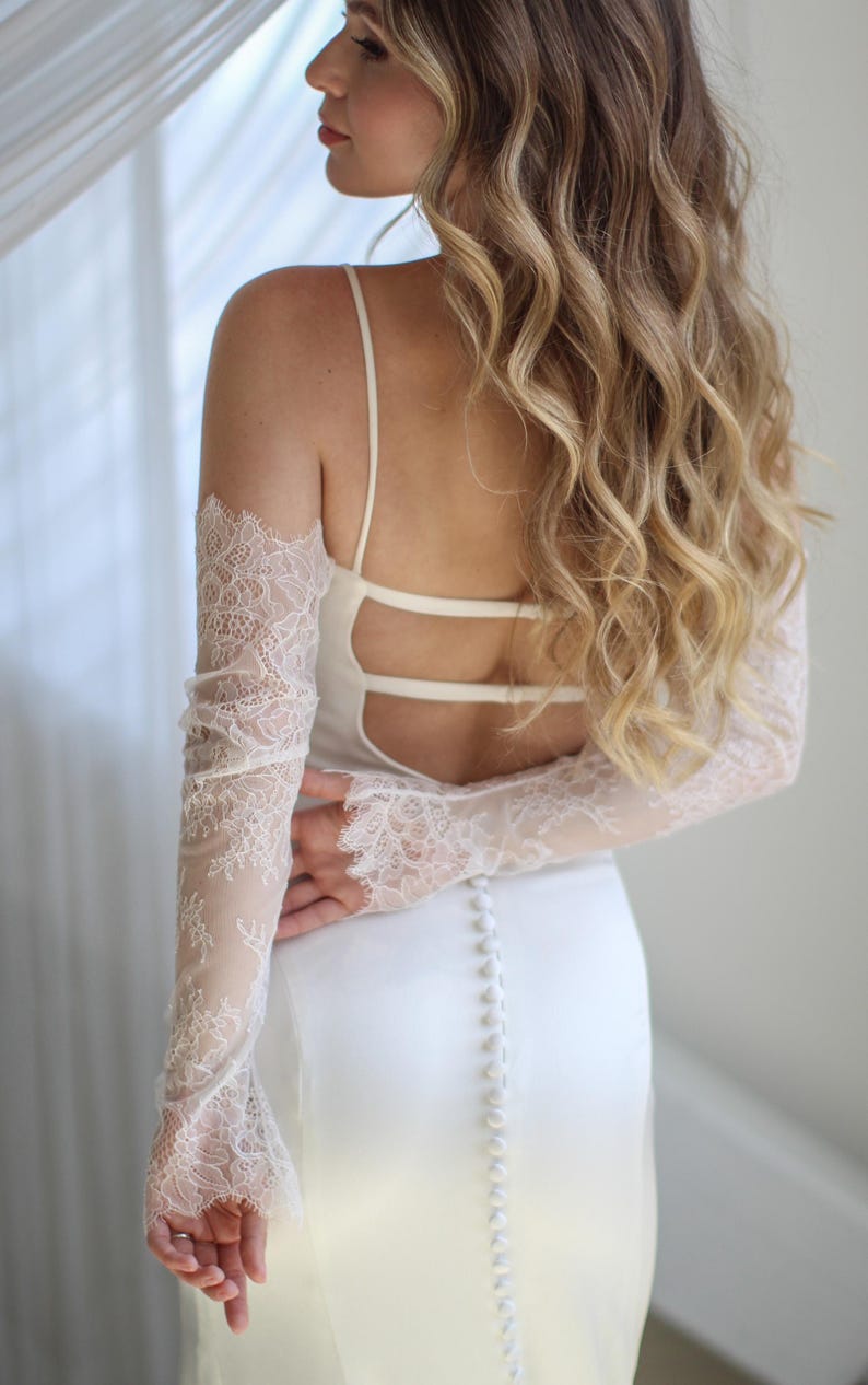 May include: A white wedding dress with a low back and spaghetti straps. The dress features a row of buttons down the back and sheer lace sleeves. The bride's hair is long and wavy.