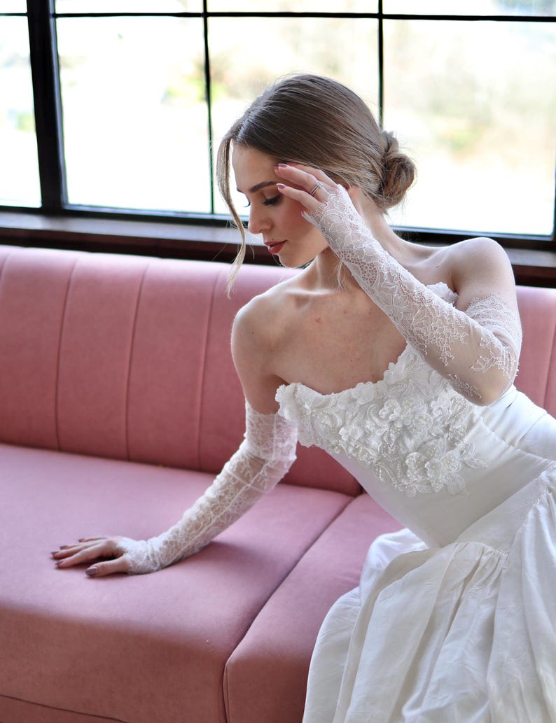May include: A white strapless wedding dress with floral embellishments and a full skirt. The bride is wearing long, delicate lace gloves. She is seated on a pink sofa near a window.