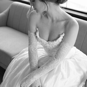 May include: A strapless white wedding dress with a full skirt. The dress features intricate lace detailing on the bodice and long, sheer lace gloves. The bride is seated on a light gray sofa.