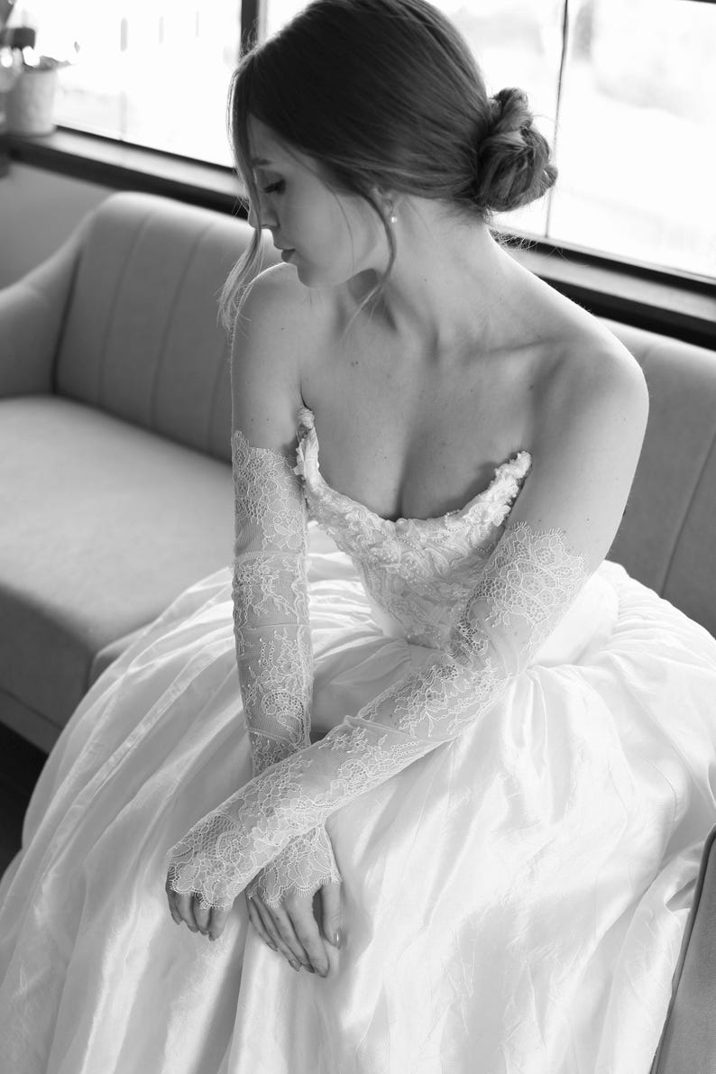 May include: A strapless white wedding dress with a full skirt. The dress features intricate lace detailing on the bodice and long, sheer lace gloves. The bride is seated on a light gray sofa.