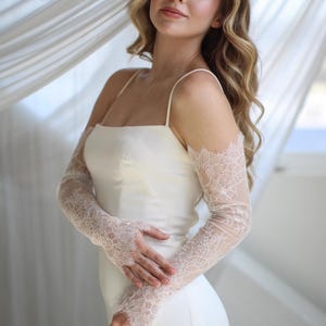 May include: A white satin wedding dress with spaghetti straps. The dress is paired with sheer, long lace sleeves. The sleeves are a light pink color and feature a delicate floral lace pattern. The background is a soft, neutral color.