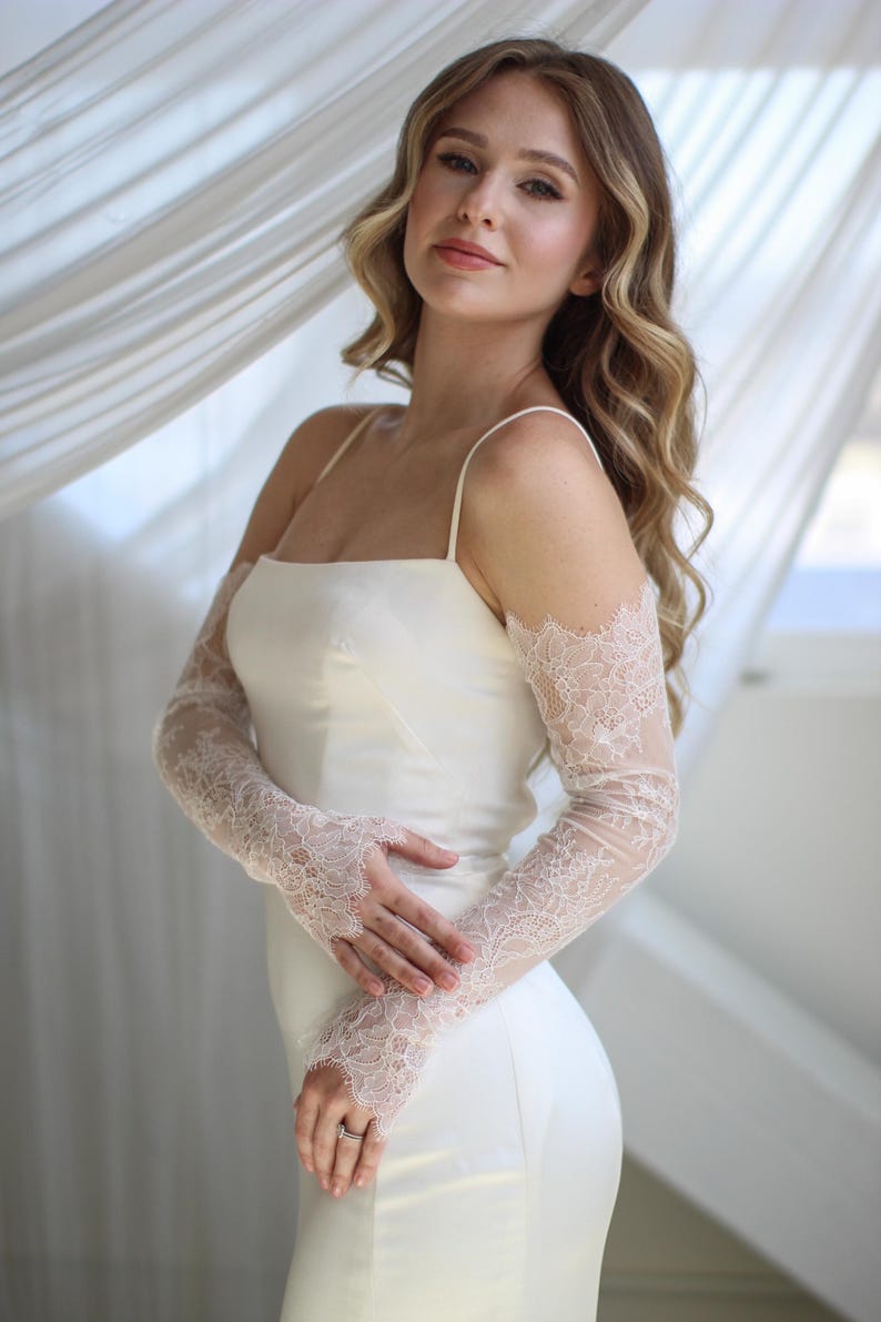 May include: A white satin wedding dress with spaghetti straps. The dress is paired with sheer, long lace sleeves. The sleeves are a light pink color and feature a delicate floral lace pattern. The background is a soft, neutral color.