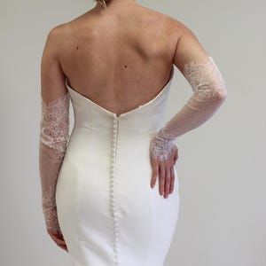 May include: A white strapless wedding dress with a fitted bodice and a flowing skirt. The dress has a row of buttons down the back and is paired with delicate lace gloves.