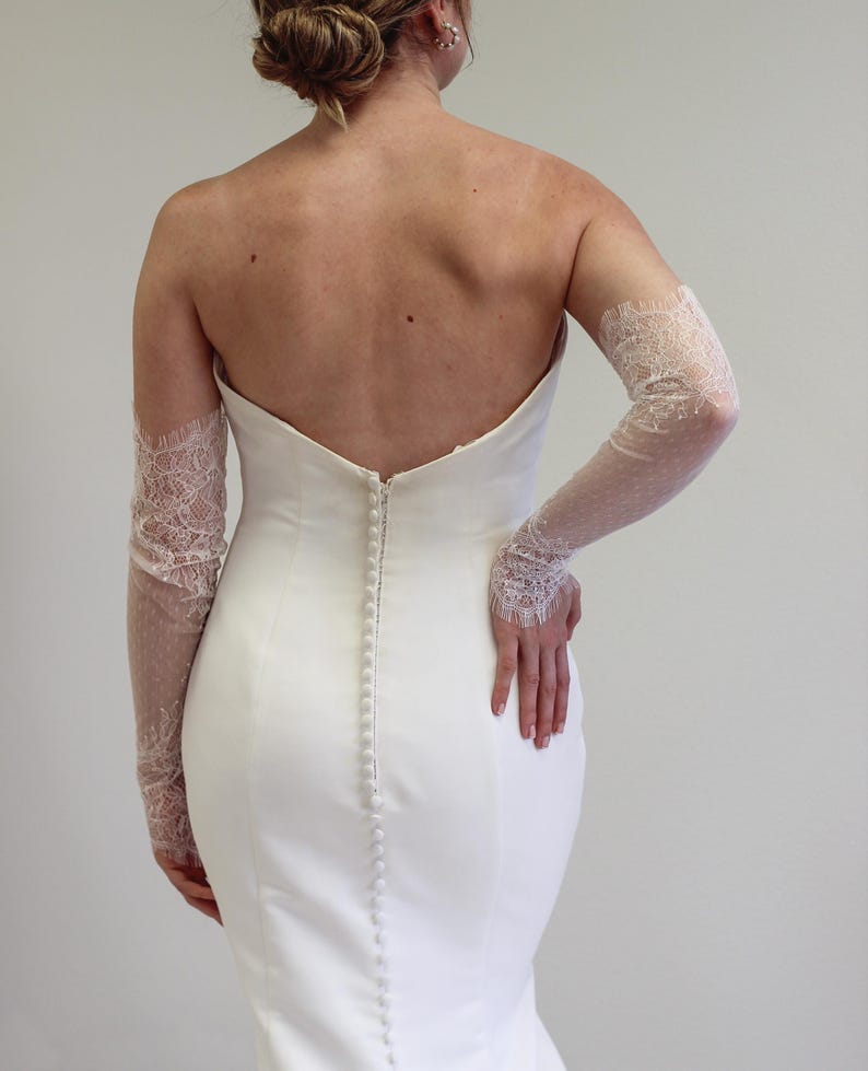 May include: A white strapless wedding dress with a fitted bodice and a flowing skirt. The dress has a row of buttons down the back and is paired with delicate lace gloves.