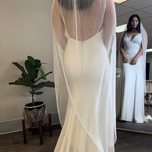 May include: A white wedding dress with a long train and a sheer veil with vertical pleats. The dress has a simple, elegant design and a fitted bodice.