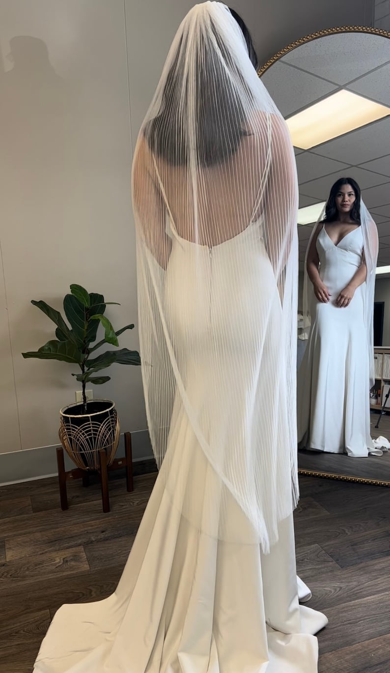 May include: A white wedding dress with a long train and a sheer veil with vertical pleats. The dress has a simple, elegant design and a fitted bodice.