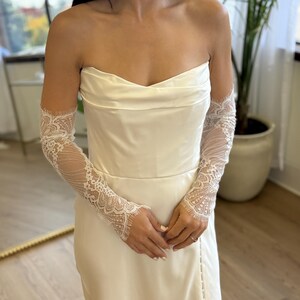 May include: Ivory strapless wedding dress with a fitted bodice and a side slit. The dress is paired with delicate, sheer lace sleeves. The sleeves feature intricate floral patterns and extend to the wrists, adding a touch of elegance.