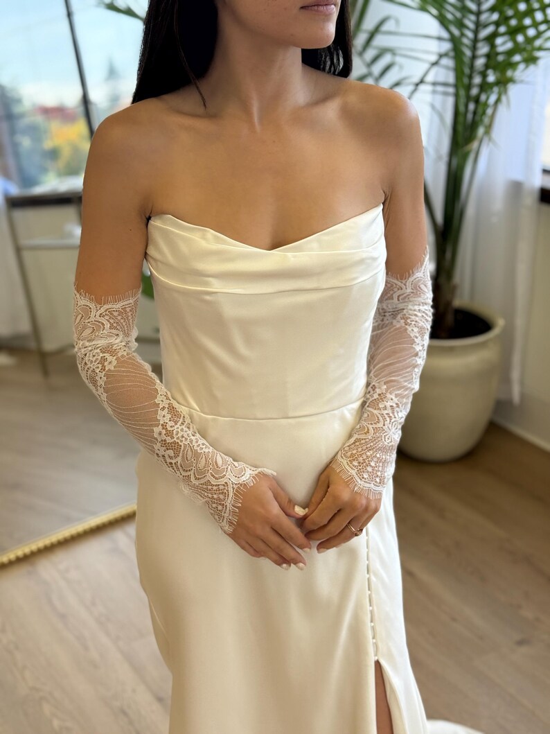 May include: Ivory strapless wedding dress with a fitted bodice and a side slit. The dress is paired with delicate, sheer lace sleeves. The sleeves feature intricate floral patterns and extend to the wrists, adding a touch of elegance.