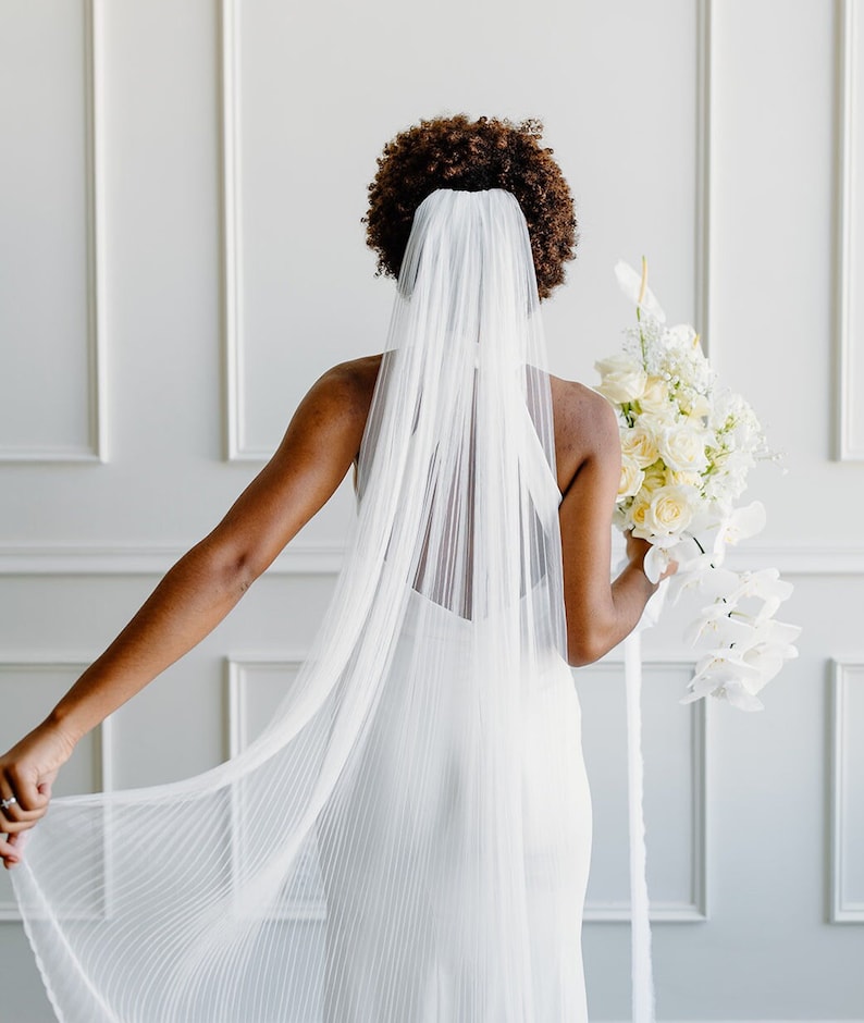 May include: A bride wearing a white wedding dress and a long, white veil. She is holding a bouquet of white flowers.