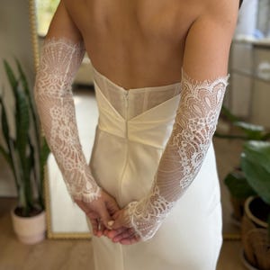 May include: A strapless ivory wedding dress with a fitted bodice and a flowing skirt. The dress is accessorized with long, delicate lace sleeves. The sleeves are intricately designed with floral patterns and scalloped edges, adding a touch of elegance.