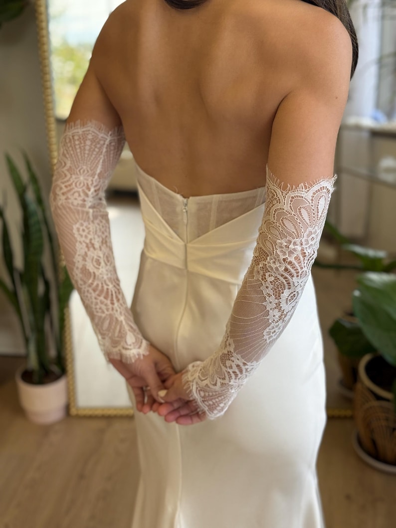 May include: A strapless ivory wedding dress with a fitted bodice and a flowing skirt. The dress is accessorized with long, delicate lace sleeves. The sleeves are intricately designed with floral patterns and scalloped edges, adding a touch of elegance.