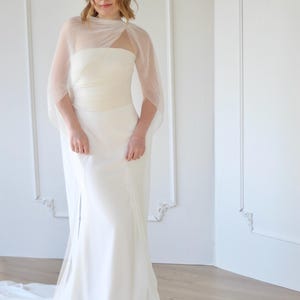 May include: A white wedding dress with a strapless bodice and a flowing skirt. The dress features a sheer, pleated cape that drapes over the shoulders. The dress is floor-length and has a subtle train. The model is wearing a bracelet.