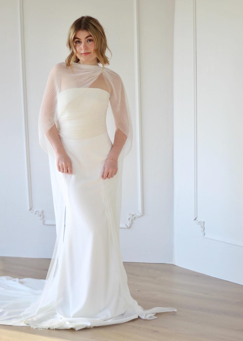 May include: A white wedding dress with a strapless bodice and a flowing skirt. The dress features a sheer, pleated cape that drapes over the shoulders. The dress is floor-length and has a subtle train. The model is wearing a bracelet.