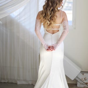 May include: A white wedding dress with a low back and a long train. The dress has a lace overlay on the sleeves and a row of buttons down the back.
