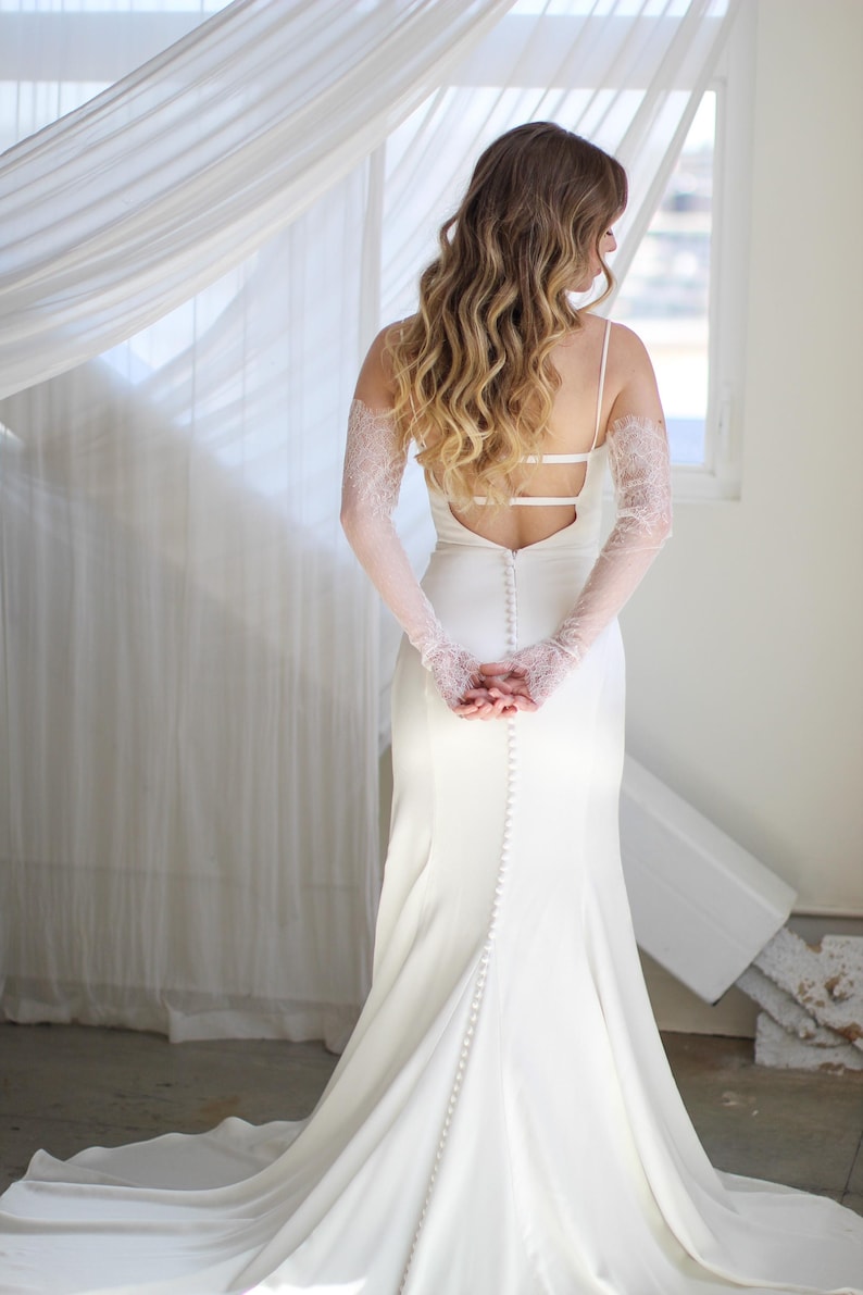 May include: A white wedding dress with a low back and a long train. The dress has a lace overlay on the sleeves and a row of buttons down the back.