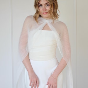 May include: A white wedding dress with a sheer, pleated cape. The dress is strapless and fitted, with a simple design. The cape is attached at the shoulders and drapes over the arms. The overall look is elegant and minimalist.