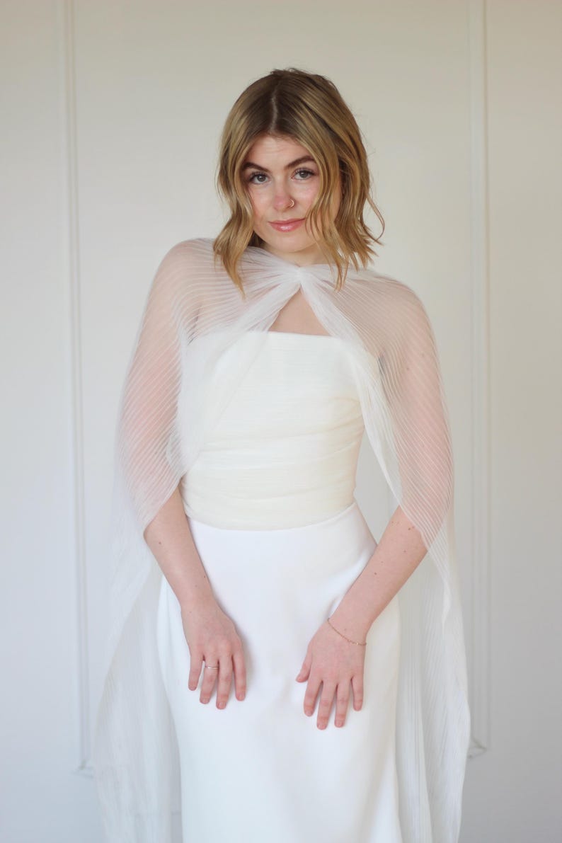 May include: A white wedding dress with a sheer, pleated cape. The dress is strapless and fitted, with a simple design. The cape is attached at the shoulders and drapes over the arms. The overall look is elegant and minimalist.