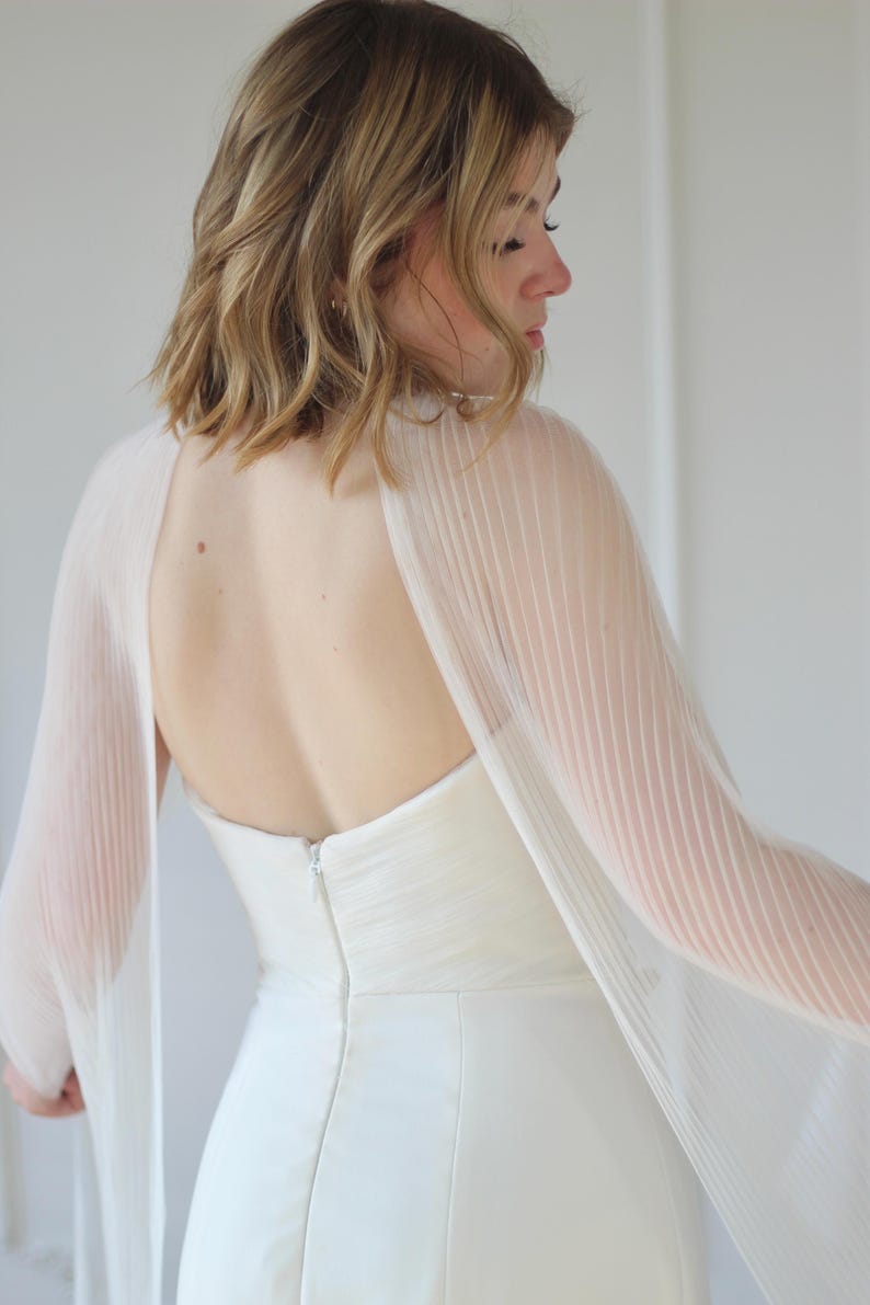 May include: A white wedding dress with a sheer, pleated cape. The dress has a fitted bodice and a simple, elegant design. The cape drapes over the shoulders and arms, adding a touch of sophistication. The back of the dress features a zipper.