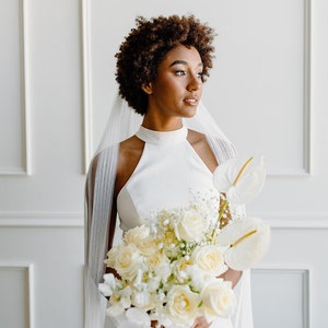 May include: A bride with short, curly hair wears a white halter-neck wedding dress and a white veil. She holds a bouquet of white roses, orchids, and other white flowers.