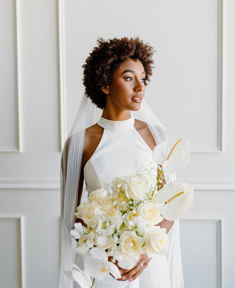 May include: A bride with short, curly hair wears a white halter-neck wedding dress and a white veil. She holds a bouquet of white roses, orchids, and other white flowers.