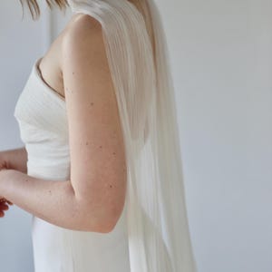 May include: A white wedding dress with a draped, flowing cape. The dress has a simple, elegant design with a strapless bodice. The cape is made of a sheer, pleated fabric that cascades down the back.