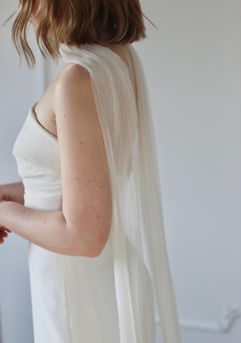 May include: A white wedding dress with a draped, flowing cape. The dress has a simple, elegant design with a strapless bodice. The cape is made of a sheer, pleated fabric that cascades down the back.