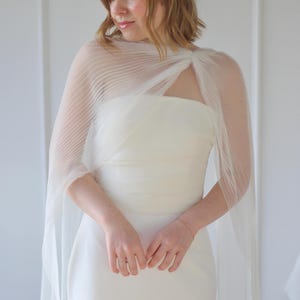May include: A white, strapless wedding dress with a sheer, pleated cape. The cape drapes over the shoulders and arms, creating a flowing silhouette. The dress is simple and elegant, perfect for a wedding or special occasion.
