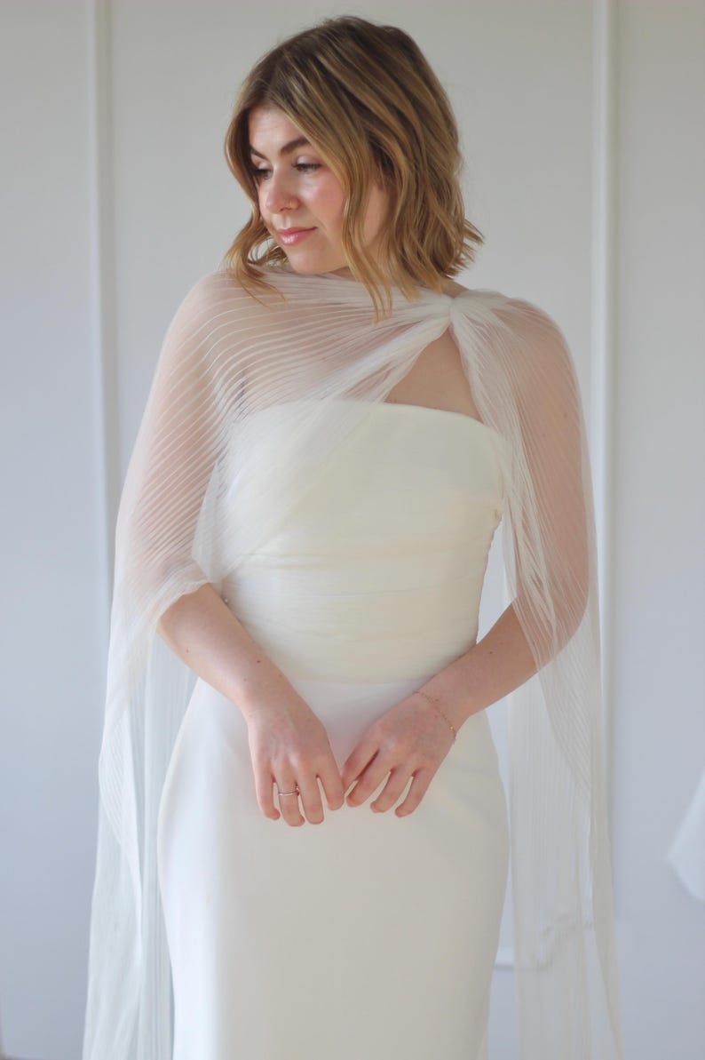 May include: A white, strapless wedding dress with a sheer, pleated cape. The cape drapes over the shoulders and arms, creating a flowing silhouette. The dress is simple and elegant, perfect for a wedding or special occasion.