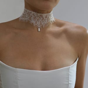 May include: A white lace choker necklace featuring an intricate floral pattern with a scalloped, fringed bottom edge. A single, small, white teardrop-shaped pendant hangs from the center of the lace band, resting against the base of the neck.