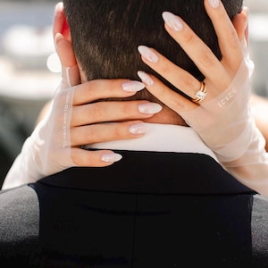 Puede incluir: Primer plano de las manos de una novia con uñas largas, ovaladas y blancas. La novia lleva un guante blanco con el texto "To Have and To Hold" en él. La novia está sujetando el cuello del novio con su otra mano, que también lleva un guante blanco. La novia lleva un anillo de oro con un gran diamante cuadrado.