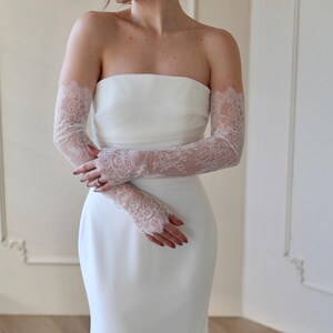 May include: A strapless white wedding dress with delicate, sheer lace gloves. The gloves extend from the wrist to the upper arm, featuring intricate floral lace patterns. The dress has a simple, elegant design.
