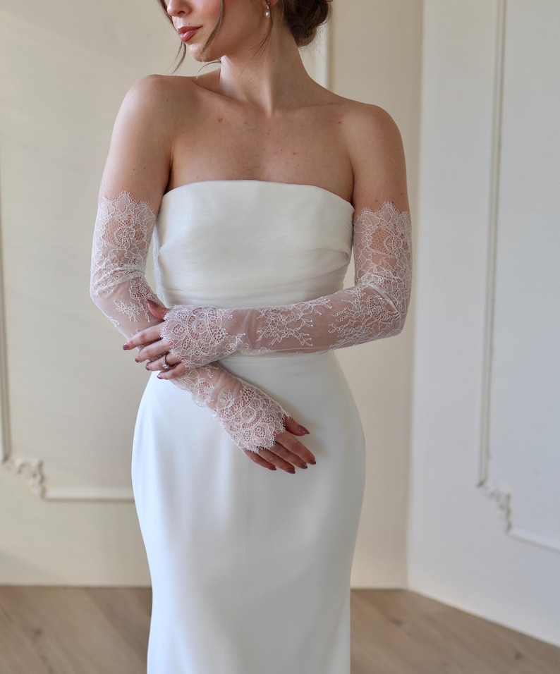 May include: A strapless white wedding dress with delicate, sheer lace gloves. The gloves extend from the wrist to the upper arm, featuring intricate floral lace patterns. The dress has a simple, elegant design.