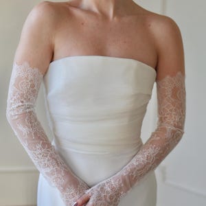 May include: A strapless white wedding dress with delicate, sheer lace gloves. The gloves extend from the wrist to the upper arm, featuring intricate floral lace patterns. The dress has a simple, elegant design.