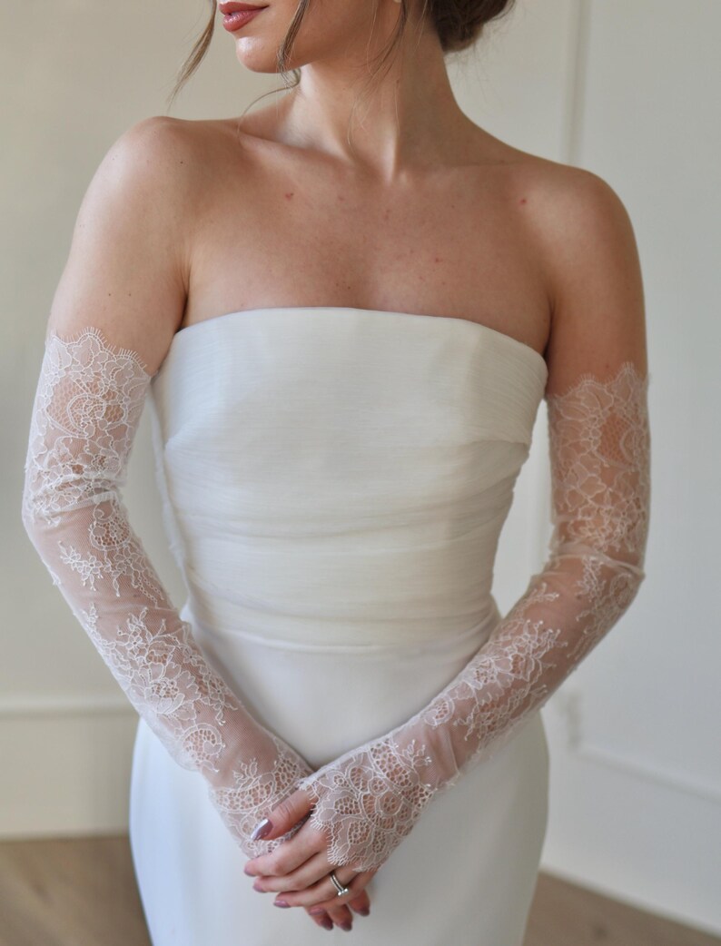 May include: A strapless white wedding dress with delicate, sheer lace gloves. The gloves extend from the wrist to the upper arm, featuring intricate floral lace patterns. The dress has a simple, elegant design.