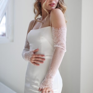May include: A bride wearing a white satin wedding dress with spaghetti straps and sheer lace gloves. The gloves are long, covering the arms to the wrists, and feature a delicate floral lace design. The background is a bright, neutral space.