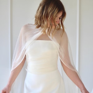 May include: A white wedding dress with a sheer, pleated cape. The dress has a simple, elegant design with a fitted bodice and a flowing skirt. The cape drapes over the shoulders and arms, adding a touch of sophistication.