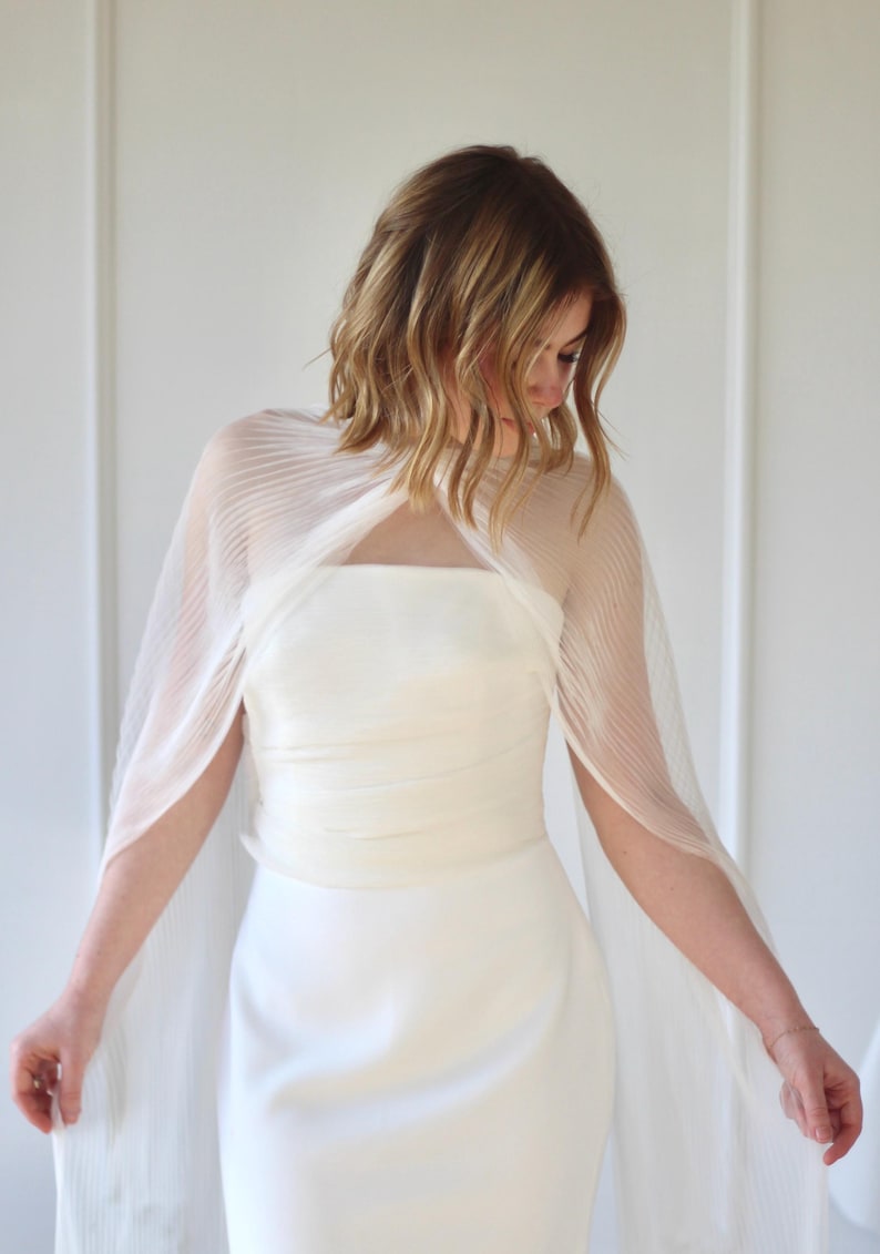 May include: A white wedding dress with a sheer, pleated cape. The dress has a simple, elegant design with a fitted bodice and a flowing skirt. The cape drapes over the shoulders and arms, adding a touch of sophistication.