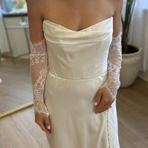 May include: A strapless ivory wedding dress with a fitted bodice and a flowing skirt. The dress features a high slit and is paired with delicate lace sleeves. The overall design is elegant and suitable for bridal wear.