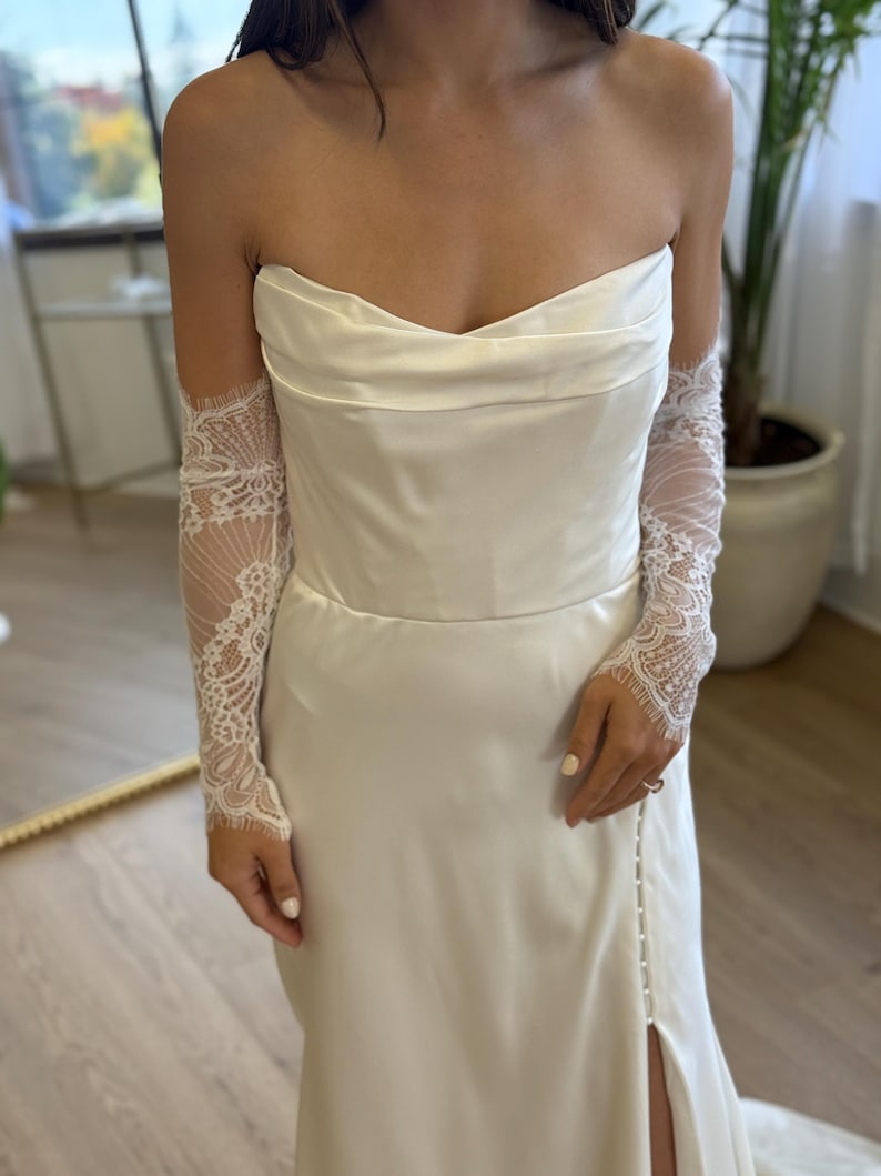 May include: A strapless ivory wedding dress with a fitted bodice and a flowing skirt. The dress features a high slit and is paired with delicate lace sleeves. The overall design is elegant and suitable for bridal wear.