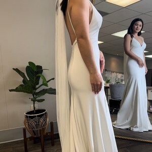 May include: A white wedding dress with a long train and a simple, elegant design. The dress has a V-neckline and a fitted bodice. The bride is wearing a white veil.
