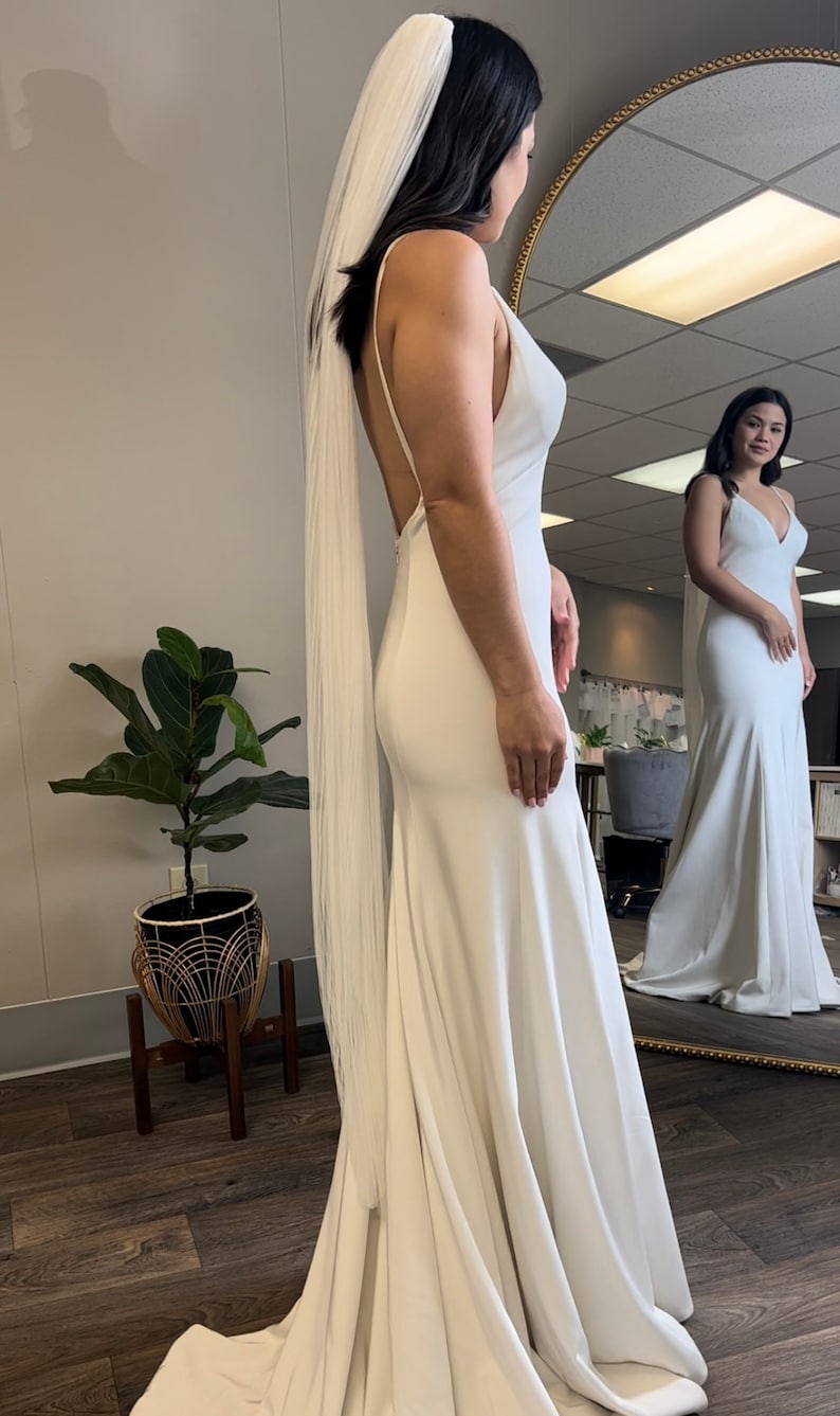 May include: A white wedding dress with a long train and a simple, elegant design. The dress has a V-neckline and a fitted bodice. The bride is wearing a white veil.