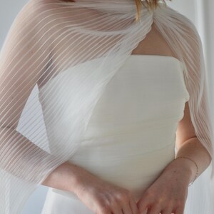 May include: A white, strapless wedding dress with a sheer, pleated wrap. The wrap drapes over the shoulders and arms. The dress is a simple, elegant design, perfect for a wedding or special occasion.